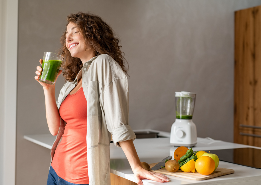 Frau trinkt grünen Smoothie in moderner Küche, frisches Obst und Mixer auf der Arbeitsfläche; gesunde Ernährung