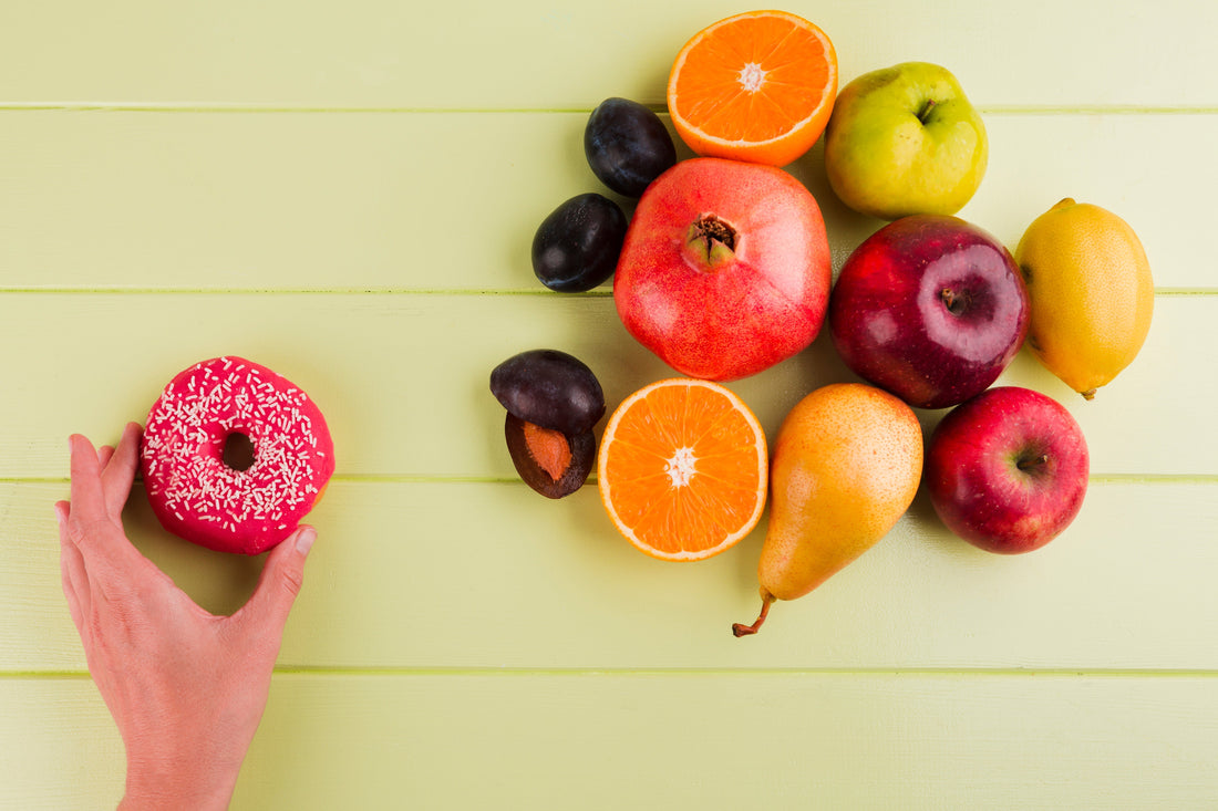 Hand greift nach pinkem Donut neben frischem Obst wie Äpfeln, Orangen und Birnen; Vergleich ungesunde und gesunde Ernährung