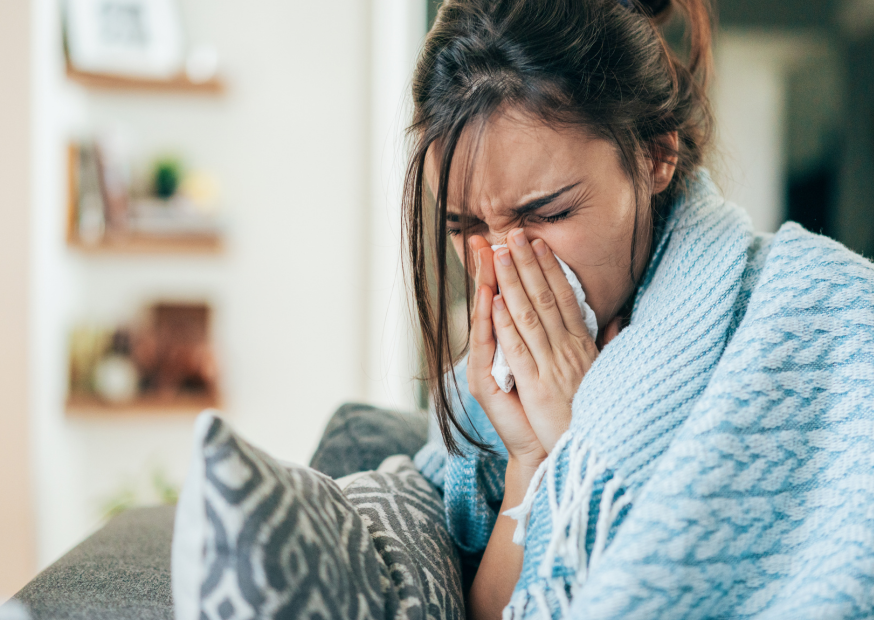 Junge Frau sitzt mit Decken eingewickelt auf einer Couch und hält sich die Hände vor das Gesicht