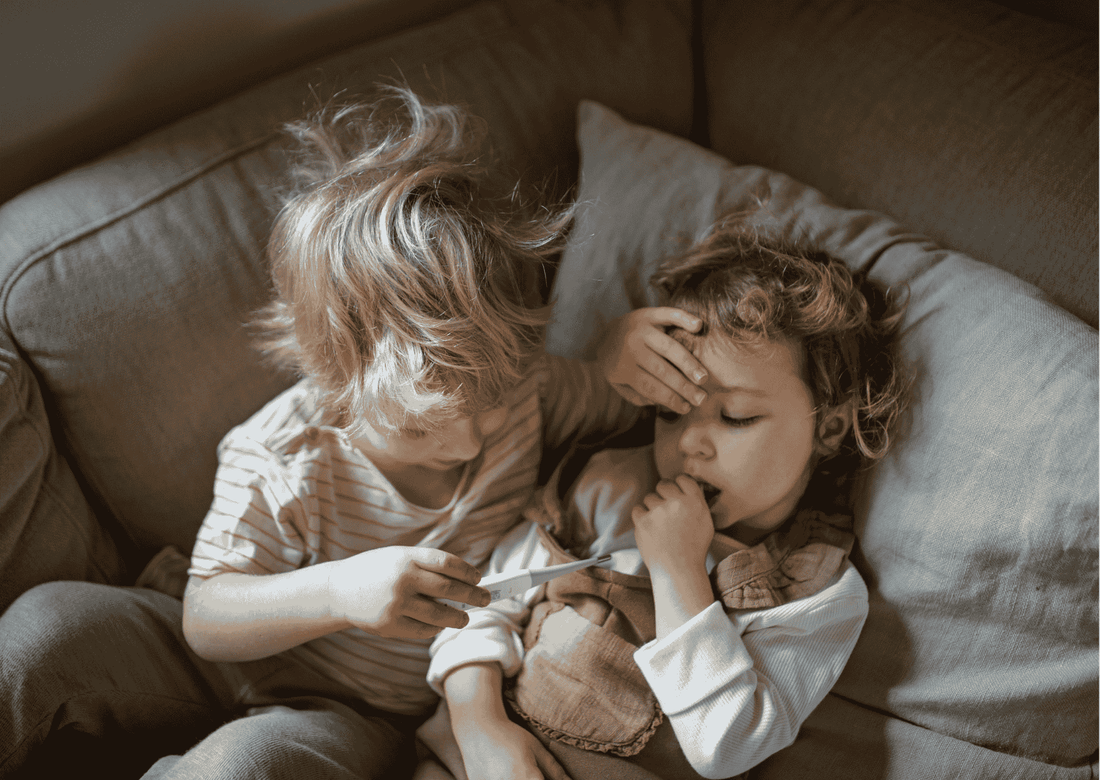Kind misst Fieber bei krankem Geschwisterkind auf Sofa, Symbol für Fürsorge