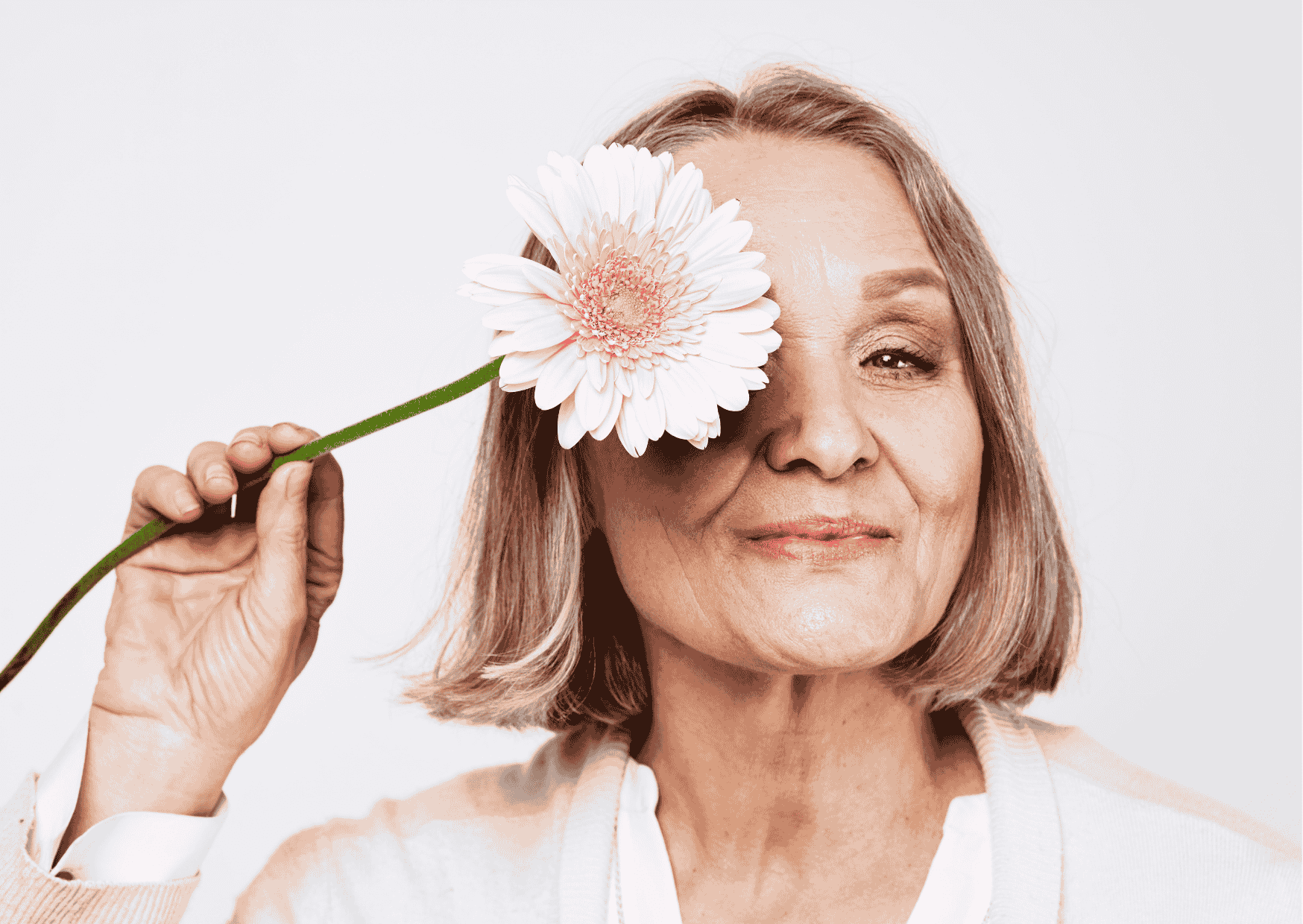 Lächelnde ältere Frau mit Kurzhaarfrisur, die eine Blume vor ihr Auge hält; natürliches Portrait, sanfter Hintergrund