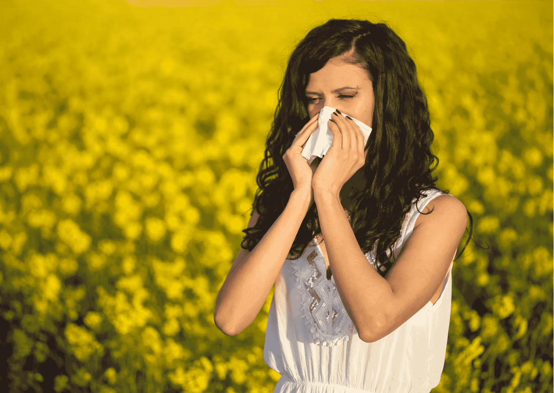Junge Frau steht im blühenden Rapsfeld und putzt sich die Nase; typische Allergiesituation im Frühling