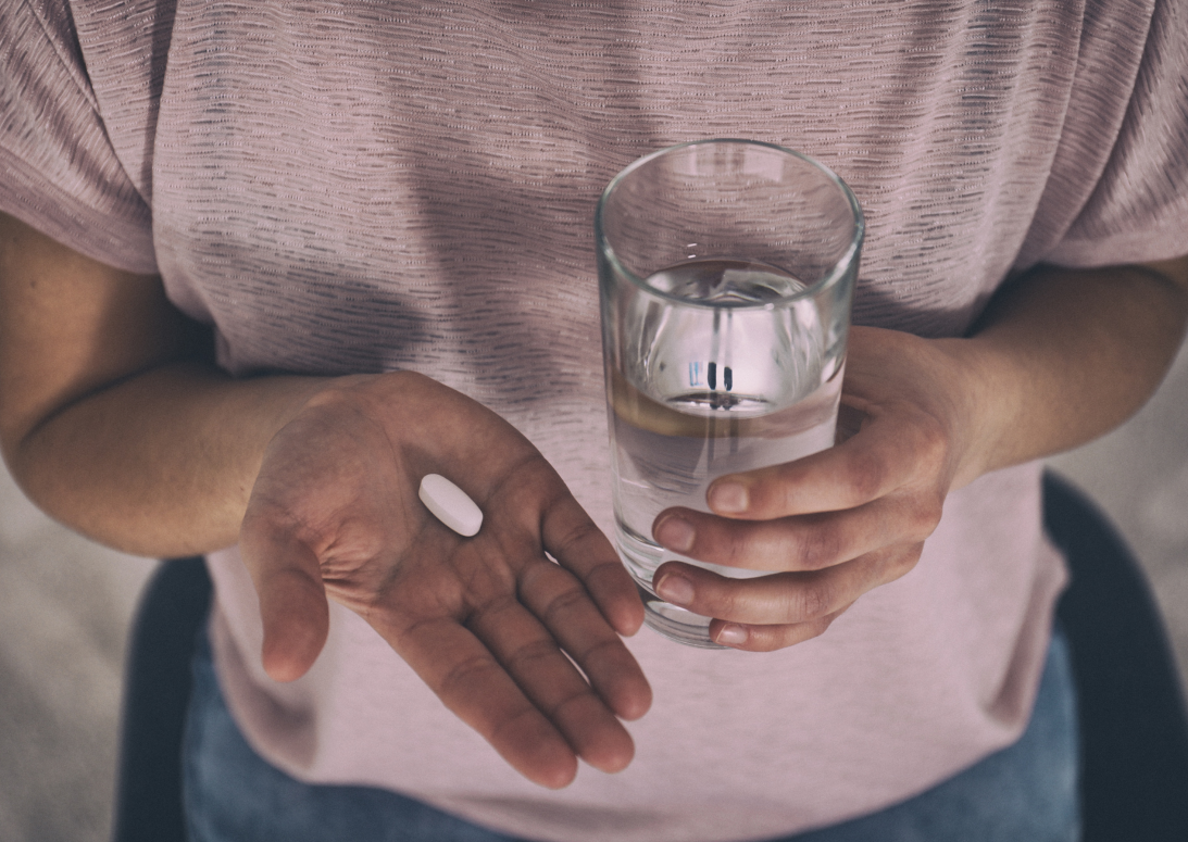 Eine Frau hält eine Pille in ihrer flachen Hand. In der anderen Hand hat sie ein Glas Wasser