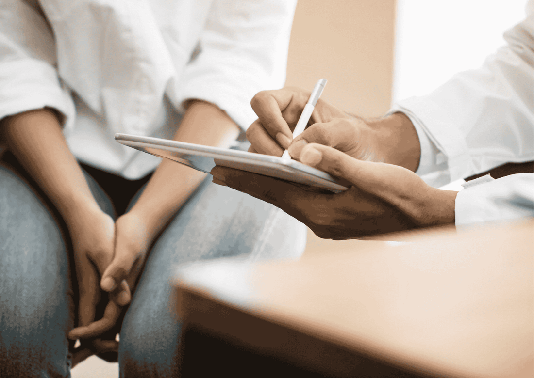 Arzt schreibt auf Tablet während Patientin mit gefalteten Händen sitzt; medizinisches Beratungsgespräch in Nahaufnahme