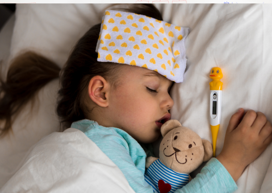Krankes Kind schläft im Bett mit Kühlpflaster auf der Stirn, Kuscheltier und Fieberthermometer daneben