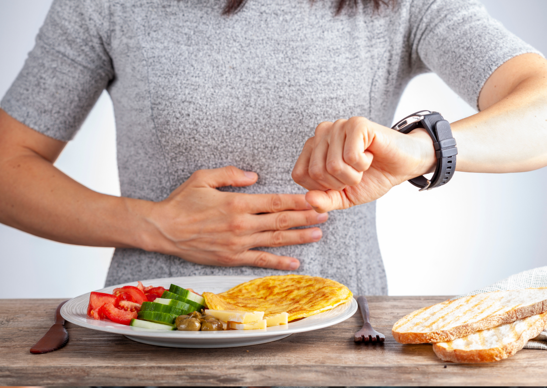 Teller mit frischen Tomaten- und Gurkenscheiben, daneben ein Omelett. Eine Frau steht hinter dem Teller, hält sich den Bauch und schaut auf ihre Armbanduhr