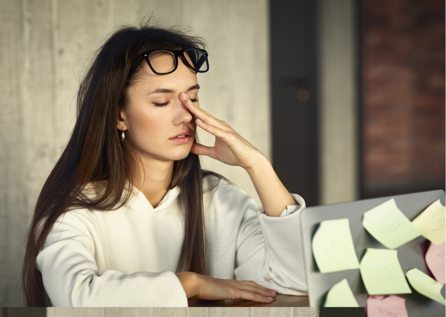Erschöpfte Frau sitzt am Schreibtisch und massiert die Stirn, Haftnotizen am Monitor; Symbol für Stress und mentale Überlastung