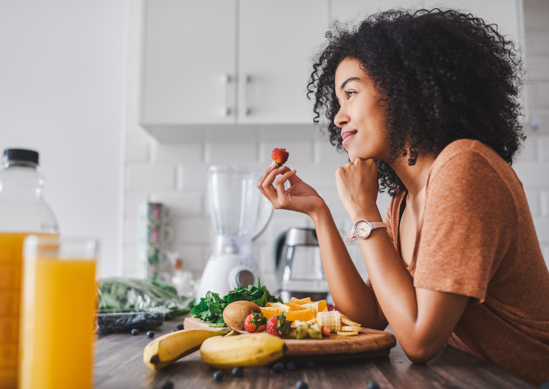 Lächelnde Frau trinkt grünen Smoothie in heller Küche, Mixer und frisches Obst stehen auf der Arbeitsfläche