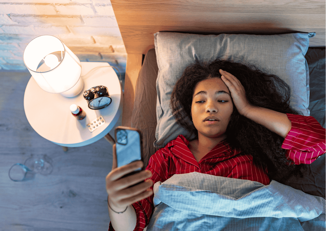 Junge Frau liegt mit Handy im Bett und hält sich den Kopf; Nachttisch mit Lampe, Uhr und Tabletten sichtbar